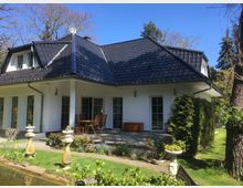 Einfamilienhaus mit weißer Fassade und dunklem Walmdach, umgeben von einem gepflegten Garten mit Rasen, Bäumen, Blumenbeeten und dekorativen Elementen wie Figuren. Eine Terrasse mit Tisch und Stühlen befindet sich an der Rückseite des Hauses.