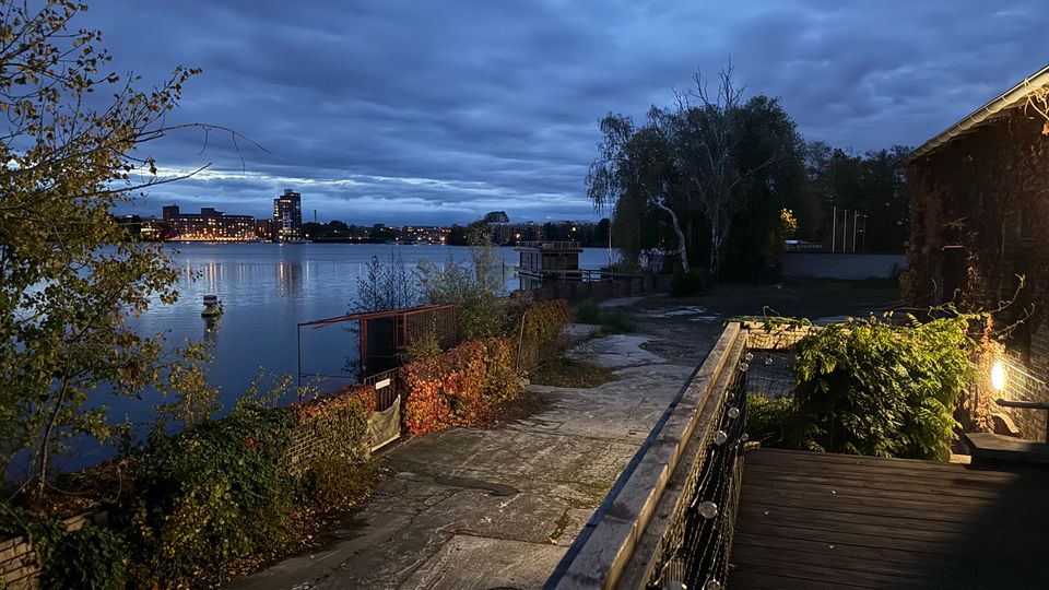 Uferpromenade an einem ruhigen Fluss bei Dämmerung, mit betoniertem Weg, Geländer und einer beleuchteten Holzterrasse neben einem Gebäude. Im Hintergrund spiegeln sich Lichter einer Stadt skyline im Wasser unter dicht bewölktem Himmel, flankiert von Bäumen und Ufervegetation.