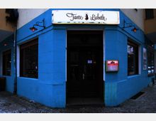 Außenansicht eines blauen Gebäudeecks mit der Eingangstür eines Lokals namens "Tante Lisbeth". Das Gebäude hat große Fenster, ein beleuchtetes Schild mit dem Schriftzug des Lokals sowie dekorative Wandlampen.