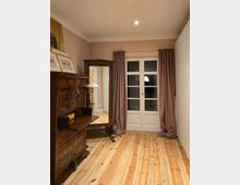 Kleines Zimmer mit hellem Holzdielenboden, beigefarbenen Wänden und einer weißen doppelflügeligen Balkontür mit braunen Vorhängen. Links stehen ein dunkler Holzsekretär und ein Spiegel-/Schrankmöbel, rechts eine schlichte weiße Wand.