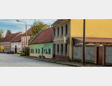 Straßenszene mit kleinen Wohnhäusern in unterschiedlichen Farben, darunter ein grünes und ein gelbes Gebäude, sowie einige Autos am Straßenrand. Der Boden besteht aus Kopfsteinpflaster, und es sind Laternen sowie eine Betonmauer mit Graffiti sichtbar.