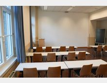 Ein Seminarraum mit mehreren Holzstühlen und weißen Tischen, die in Reihen angeordnet sind. Im Hintergrund stehen ein Rednerpult und ein großer Bildschirm, links befindet sich ein Fenster mit blauen Vorhängen.
