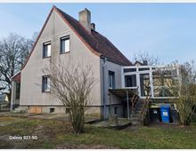 Einfamilienhaus mit Satteldach und hellem Putz, seitlich ein angebauter Wintergarten mit Glasfenstern. Vor dem Haus befinden sich eine Treppe, Mülltonnen und ein kahler Strauch, umgeben von einer Wiese und Bäumen im Hintergrund.