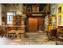 Holzwerkstatt mit rustikalem Ambiente, ausgestattet mit einer Werkbank, Werkzeugregalen, Holzschränken und gelben Fenstern an der rechten Wand. Im Hintergrund befinden sich ein Hochregal mit Stühlen und antiken Möbelstücken sowie verschiedene Arbeitsmaterialien.