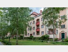 Mehrfamilienhaus im Stil der Zwischenkriegsmoderne mit hellen Fassaden, roten Fensterrahmen und kleinen Balkonen, umgeben von einer grünen Gartenanlage mit Bäumen und Rasenflächen. Im Vordergrund führt ein Gehweg an einer Straßenlaterne entlang.