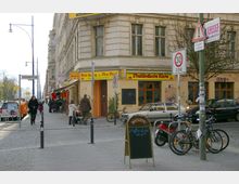 Straßenszene an der Ecke eines Altbaus mit einem asiatischen Bistro und der Aufschrift „Thailändische Küche“ auf gelber Markise. Vor dem Gebäude stehen Fahrräder, ein geparktes Auto, ein Werbeschild sowie verschiedene Verkehrsschilder, während Passanten auf dem Gehweg unterwegs sind.