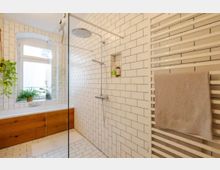 Modernes Badezimmer mit weißen U-Bahn-Fliesen an Wänden und Boden, einer bodengleichen Dusche mit Glastrennwand, einer Badewanne mit Holzverkleidung und einem großen Fenster. Ein Handtuch hängt an einem weißen Handtuchheizkörper, und Pflanzen stehen neben der Wanne.