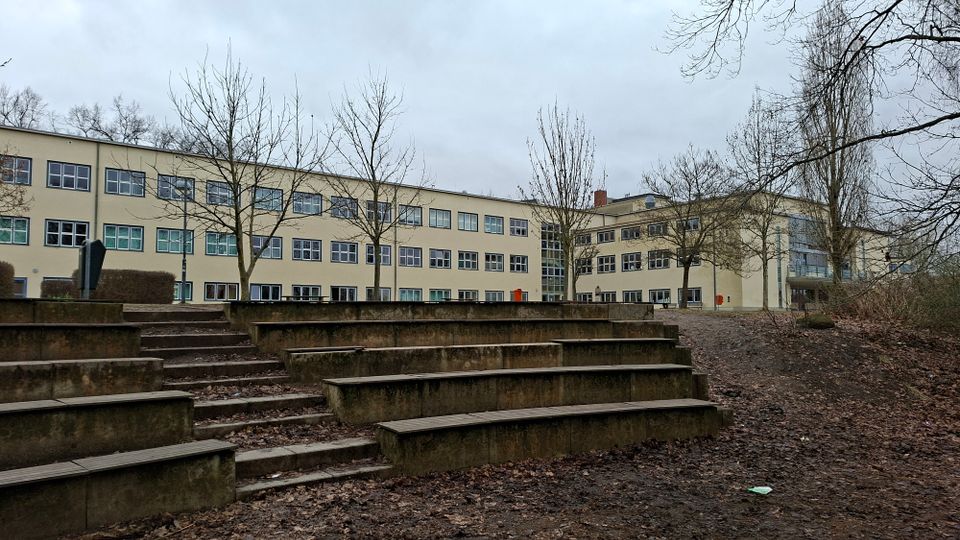 Großer, dreigeschossiger Schul- oder Verwaltungsbau mit heller Fassade und vielen rechteckigen Fenstern, im Hintergrund eines Innenhofs. Im Vordergrund stehen terrassenförmige Betonstufen wie eine kleine Freilufttribüne, umgeben von kahlen Bäumen und unbefestigtem Boden.