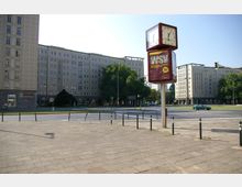 Großer Platz mit breiter Straße und typischen Gebäuden im Baustil der Nachkriegszeit. Im Vordergrund eine quadratische Uhr auf einem Pfeiler, daneben Gehwegbegrenzungen und eine Grünfläche mit Bäumen.