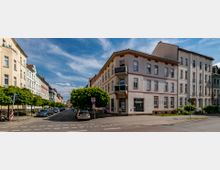 Straßenszene in einem Wohnviertel mit mehrstöckigen Altbauhäusern in hellen Farben, teilweise mit Balkonen. Entlang der Straße stehen geparkte Autos, daneben kleine Bäume, und der Himmel ist leicht bewölkt.