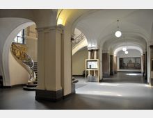 Eingangsbereich eines historischen Gebäudes mit hohen Decken und gewölbten Bögen. Links eine geschwungene Treppe mit verziertem Geländer, rechts Steinsäulen und Informationstafeln, im Hintergrund ein großes Gemälde an der Wand.