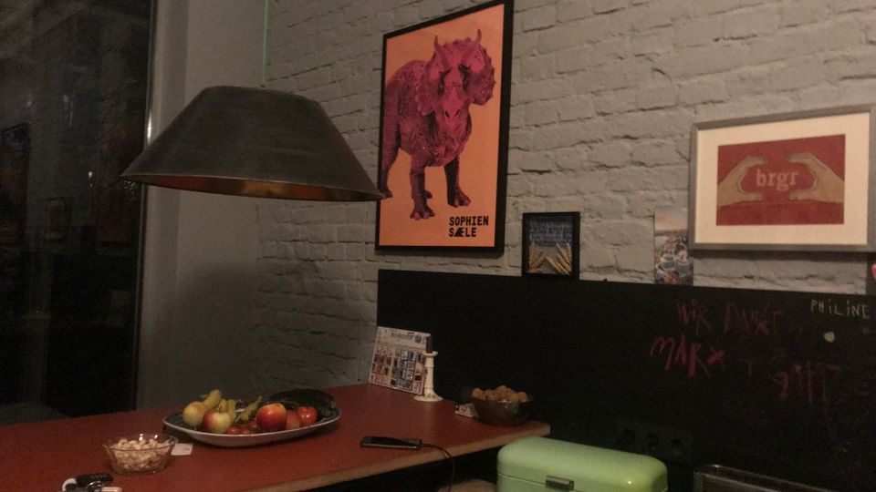 Innenraum mit grauer Backsteinwand und langen roten Tischflächen, darüber mehrere gerahmte Poster. Links hängt eine große schwarze Pendelleuchte, auf dem Tisch stehen Obstschale und Kleinteile; im Hintergrund ist eine schwarze Wandtafel und ein mintgrüner Kasten zu sehen.