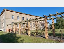 Ein historisches Gebäude mit klassischer Fassade aus Putz und Fenstern mit weißen Rahmen. Im Vordergrund eine Pergola aus Backsteinsäulen und Holzstreben, umrankt von dünnen Pflanzen, dahinter eine Rasenfläche und Bäume.