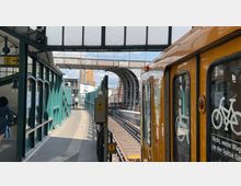 Eine oberirdische U-Bahn-Station mit grünem Stahlgeländer, transparentem Dach und einer gelben U-Bahn, die am Bahnsteig hält. Im Hintergrund sind ein gebogener Übergang, Gleise und moderne Gebäude zu sehen.