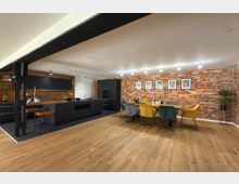 Moderne Wohnküche mit offenem Grundriss, bestehend aus einer schwarzen Einbauküche mit Holz-Arbeitsfläche links und einem Essbereich mit farbigen Polsterstühlen rechts. Die Rückwand ist aus roten Ziegelsteinen, dekoriert mit gerahmten Bildern, und der Boden besteht aus dunklem Holzparkett.