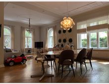 Wohnzimmer mit einem Esstisch und gepolsterten Stühlen in einem eleganten, hellen Raum mit hohen Decken und verzierten Stuckelementen. Große Fenster mit Blick ins Grüne, ein Kronleuchter sowie moderne Beleuchtung und ein kleines rotes Spielzeugauto auf dem Holzboden runden die Szene ab.