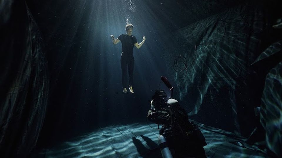Underwater cavern-like space with dark rock walls and a flat floor, lit by sunbeams and rippling light patterns from the surface above. A scuba diver with equipment is near the bottom, with floating particles visible in the water.