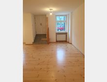 Heller Innenraum mit Holzfußboden, einer Treppe mit Fliesenstufen, die zu einer weißen Tür führt, sowie einem Fenster mit Heizkörper darunter. Draußen vor dem Fenster ist ein rotes Auto sichtbar.