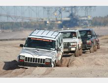 Geländefahrzeuge fahren hintereinander durch eine sandige, unebene Offroad-Strecke. Im Hintergrund ist eine Industriekulisse mit einer großen Förderanlage und Bäumen am Horizont zu sehen.