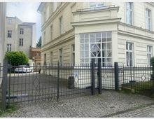 Eingezäunter Vorhof eines hellen historischen Gebäudes mit großen, weißen Fenstern, die symmetrische Gitterdetails aufweisen. Im Hintergrund befinden sich ein gepflasterter Weg, geparkte Autos und angrenzende Gebäude.