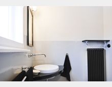 Modernes Badezimmer mit einem weißen Wandwaschbecken auf einer dunklen Arbeitsfläche, einer Wandarmatur aus Edelstahl und einem rechteckigen Spiegel. An der Wand befinden sich ein schwarzer Handtuchhalter, ein schwarzer Heizkörper und eine kleine Ablage.