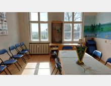 Ein heller Raum mit großen Fenstern, die auf eine ländliche Landschaft blicken, enthält eine lange Tischgruppe mit Stühlen sowie einen Schrank und gestapelte Klappstühle an der Seite. Auf einer Wand ist ein Wandgemälde mit einer Naturszene zu sehen, und ein Banner auf dem Tisch trägt die Aufschrift „DAV Ortsgruppe Viernaden 2001 e.V.“.