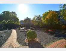 Gartenbereich mit gepflastertem Weg, Kiesflächen und mehreren Tischen und Stühlen aus Holz und Metall. Umgeben von Bäumen mit herbstlich gefärbtem Laub, im Hintergrund Gebäude und blauer Himmel.