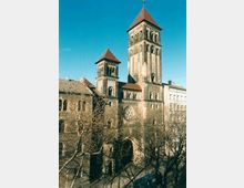 Historische Kirche mit zwei hohen Türmen und roten Spitzdächern in romanischem Stil, umgeben von kahlen Bäumen; im Vordergrund eine Straße mit einem blauen Fahrzeug.