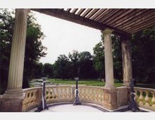 Pergola mit steinernen Säulen und Geländer, die eine Aussicht auf einen Park mit Teich und umliegenden Bäumen bieten. Der Boden ist mit hellen Fliesen gestaltet, und dekorative Metallständer sind entlang des Geländers positioniert.