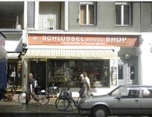 Fassade eines Schlüsseldienstladens mit orangefarbenem Schild, auf dem „Schlüssel Service Shop“ steht. Im Schaufenster sind Schlüssel und Geräte ausgestellt, während Passanten mit Fahrrädern und ein geparktes Auto im Vordergrund sichtbar sind.
