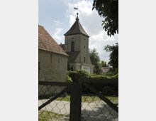 Steinerne Kirche mit rechteckigem Turm und spitzem Ziegeldach, gekrönt von einer Wetterfahne, umgeben von Efeu und Bäumen; im Vordergrund ein Metallzaun.
