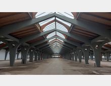 Großer, leerer Innenraum einer Halle mit einer Dachkonstruktion aus Holz und Metallträgern. Das Dach ist teilweise verglast und lässt Tageslicht einfallen, während die Wände weiß und schlicht gehalten sind.