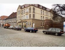 Straßenszene mit Kopfsteinpflaster, einer historischen Häuserzeile mit Fachwerk- und Jugendstilgebäuden sowie geparkten Autos. Im Hintergrund ist eine Backsteinmauer sichtbar, die an eine ältere Stadtbefestigung erinnern könnte.