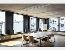 Moderner Besprechungsraum mit großen, bodentiefen Fenstern, die viel Tageslicht hereinlassen. Der Raum verfügt über einen rechteckigen Tisch mit Holzstühlen, einen Flipchart und einen Holzboden; im Hintergrund sind Außentreppen und ein Bürogebäude sichtbar.