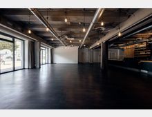 Großer, leerer Innenraum mit Betonwänden und -decke, sichtbaren Lüftungsrohren und freihängenden Glühbirnen. Auf der linken Seite befinden sich große Glastüren mit Blick nach draußen, rechts ein Tresen aus dunklem Holz.