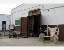 Eingang zu einem einfachen, braun-weiß gestrichenen landwirtschaftlichen Gebäude mit geöffneter Holztür. Vor dem Gebäude stehen eine Schubkarre, ein Hufschuhwagen und eine Bank, im Inneren sind Eimer und Werkzeuge erkennbar.