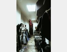 Narrow backstage dressing room or storage corridor with fluorescent ceiling lights, wood plank flooring, and white walls. Clothing and coats hang along the right side, while instrument cases, a mirror and shelf, and scattered gear line the left, leading to a door at the far end.