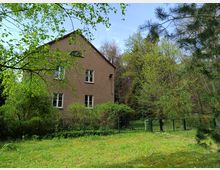 Zweistöckiges Wohnhaus mit brauner Fassade und weißen Fensterrahmen, umgeben von üppigem Grün und Bäumen. Im Vordergrund befindet sich ein Gartengrundstück mit Rasenfläche und einem schwarzen Metallzaun.