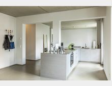 Moderne, minimalistisch eingerichtete Küche mit weißer Einbauküche in L-Form und einer zentralen Kücheninsel aus Betonoptik. An der Wand links hängt ein bunter Garderobenhaken, und im Hintergrund sind ein Spiegel und schlicht gehaltene Dekorationen sichtbar.