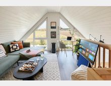 Helles Wohnzimmer in einem Dachgeschoss mit schrägen weißen Holzdecken, großen bodentiefen Fenstern und ländlichem Ausblick. Der Raum ist modern eingerichtet mit einer grünen Eckcouch, einem ovalen Couchtisch, einem Schreibtisch mit Laptop und einem schräg stehenden Flachbildfernseher.