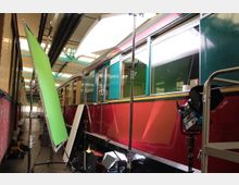 Ein roter und grüner Oldtimer-Zug steht in einer Halle mit offenen Türen, umgeben von Film- oder Fotoausrüstung wie Scheinwerfern, einem grünen Hintergrund und Stativen. Die Halle hat eine helle Decke mit sichtbaren Rohrleitungen.