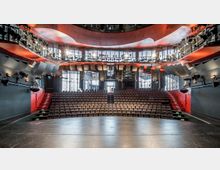 Ein moderner Theatersaal mit ansteigenden Sitzreihen, einer großen Bühne im Vordergrund und roten Akzenten an den Wänden und Treppen. Im Hintergrund sind Glaswände und eine Tür, die Tageslicht hereinlassen, sowie zahlreiche Scheinwerfer und Akustikpaneele an der Decke und den Seitenwänden.