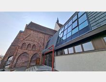 Außenansicht eines historischen Backsteingebäudes mit hohen, zugemauerten Rundbögen und einem spitzen Turm im Hintergrund. Daneben steht ein moderner Anbau mit großer Glasfassade, Metallverkleidung und einem Eingang über eine kleine Treppe.
