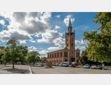 Backsteinkirche mit hohen Rundbogenfenstern und einem Glockenturm mit spitzem grünen Dach, umgeben von einem Vorplatz mit parkenden Autos und Bäumen; der Himmel ist teils bewölkt.
