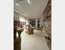 Heller, moderner Laden- oder Galerieraum mit weißem Boden, Tracklights an der Decke und großen Fenstern sowie einer Glastür zum Außenbereich. Im Raum stehen Baumstamm-Tische und Vasen mit Blumen, an der Wand hängen farbige Kunstbilder und rechts befinden sich Regale mit ausliegenden Produkten.