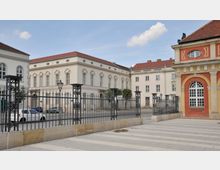 Ansicht eines historischen Platzes mit klassizistischen Gebäuden. Im Vordergrund befindet sich ein schmiedeeisernes Gitter, dahinter stehen beige-weiße und ein rot-ockerfarbenes Gebäude mit Säulen, Rundbogenfenstern und Zierelementen.