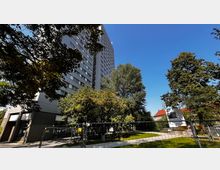 Hochhaus aus Beton und Glas mit zahlreichen Fenstern, umgeben von Bäumen und Grünflächen bei klarem Himmel; im Vordergrund ist ein Bauzaun sichtbar.
