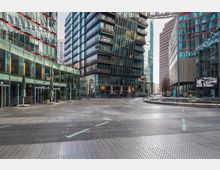 Moderner Platz mit umliegenden Gebäuden aus Glas und Metall im Sony Center, Berlin. Die Architektur zeichnet sich durch vertikale und geneigte Glasfassaden aus, mit Geschäften und Restaurants im Erdgeschoss und einer überdachten Passage oberhalb des Platzes.