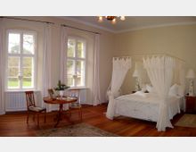 Helles Schlafzimmer mit großen Fenstern, weißen Vorhängen und einem Himmelbett mit weißen Stoffvorhängen, dekoriert mit Holzmöbeln und einem kleinen runden Tisch. Der Raum hat einen Holzboden, helle Wände und Ausblick ins Grüne.