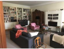 Wohnzimmer mit einer großen Bücherwand, einem Kamin und klassischen Möbeln. Der Raum enthält zwei pinkfarbene Sessel, ein großes dunkles Sofa, einen Fernseher und einen Glastisch mit dekorativen Objekten.